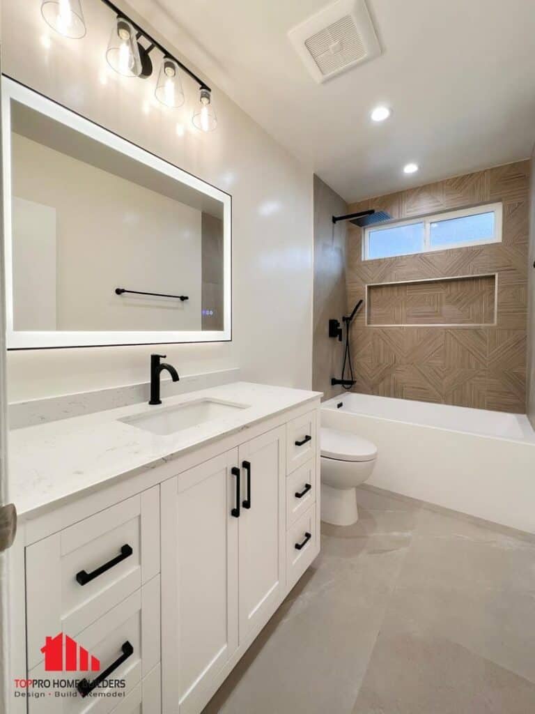 Image of a modern bathroom with a white vanity, integrated mirror lighting, and geometric tile design around the bathtub.